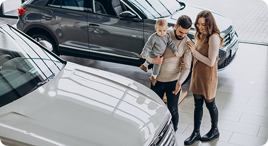 Uma família observa carros em um showroom iluminado, com uma criança no colo de um adulto perto de dois veículos estacionados.