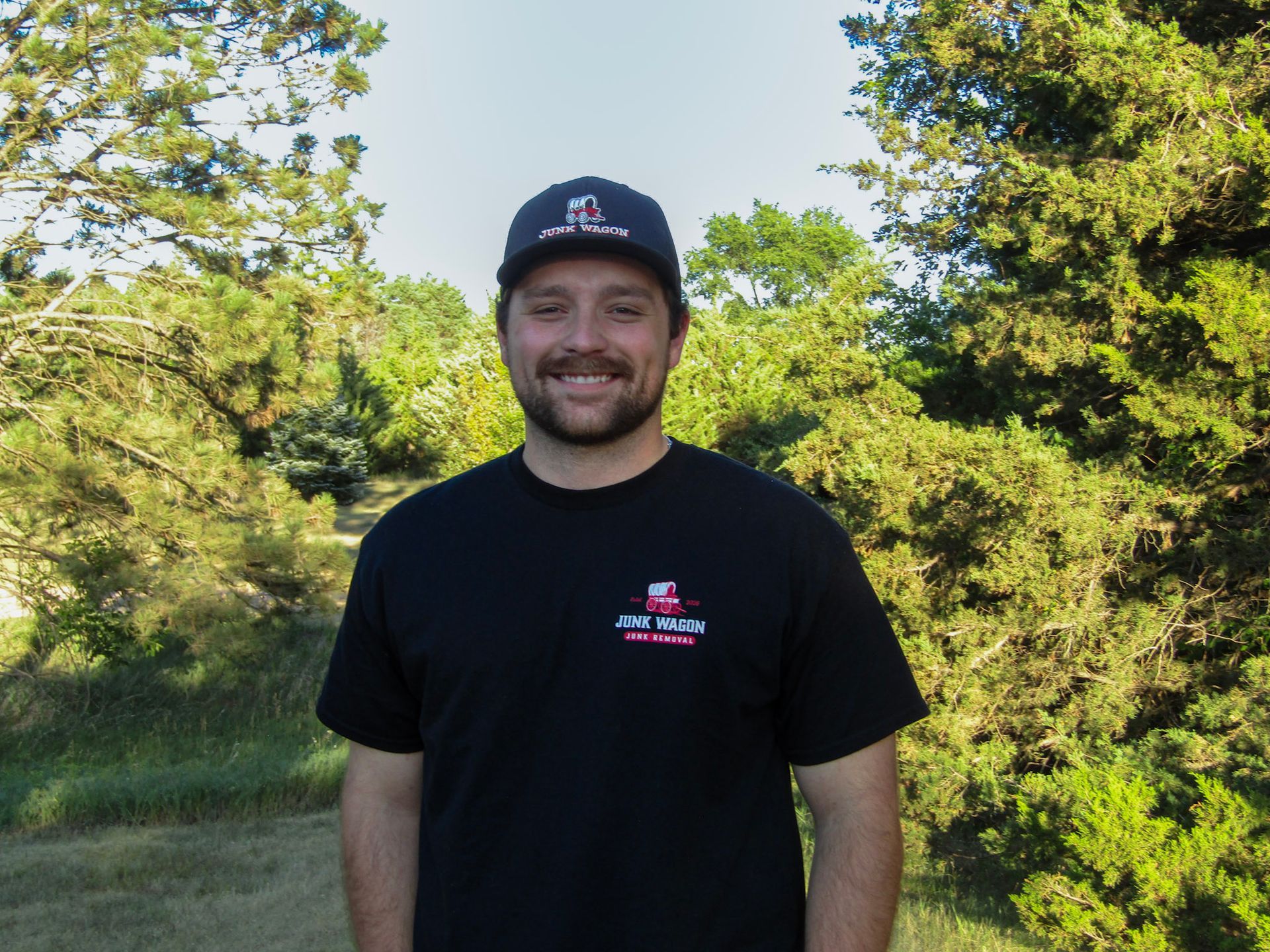 Smiling man in black T-shirt and cap standing outdoors among green trees