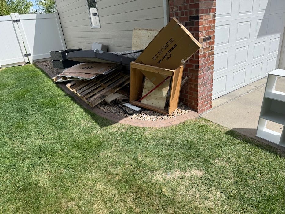 Piled wooden boards and a broken cardboard structure beside a garage on a lawn