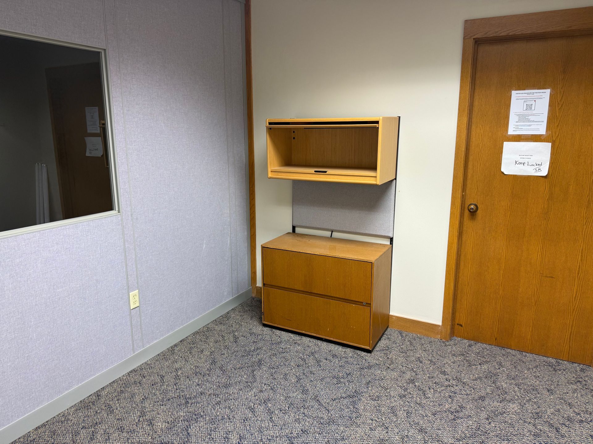 Empty office corner with gray carpet, wall cabinet, and wooden door with posted notices.