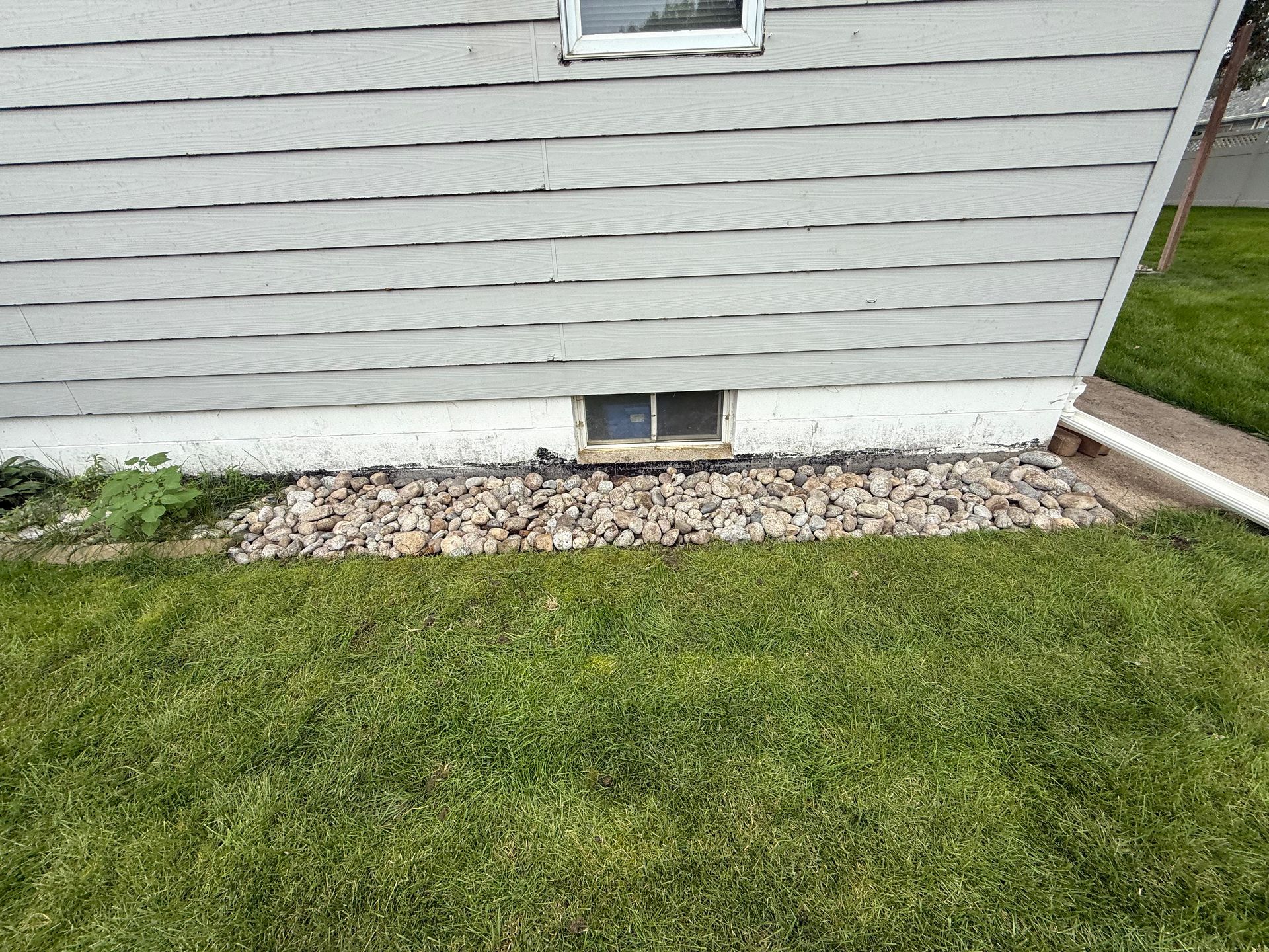House siding with a small basement window, bordered by gravel and green lawn.