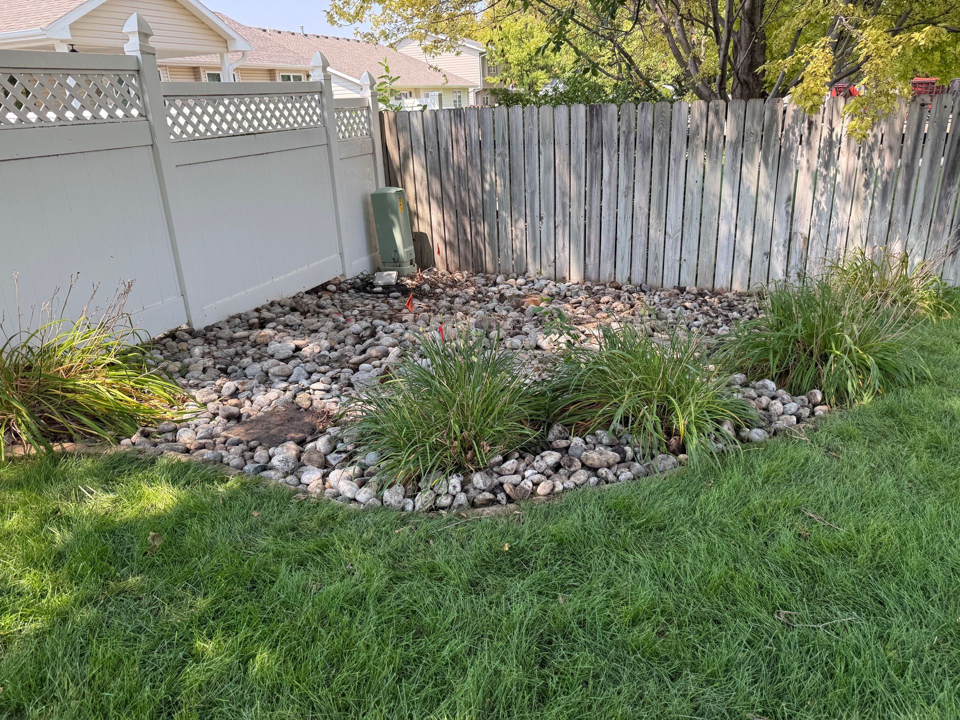 Small backyard rock garden with plants, grass, a white fence, and a weathered wooden fence.