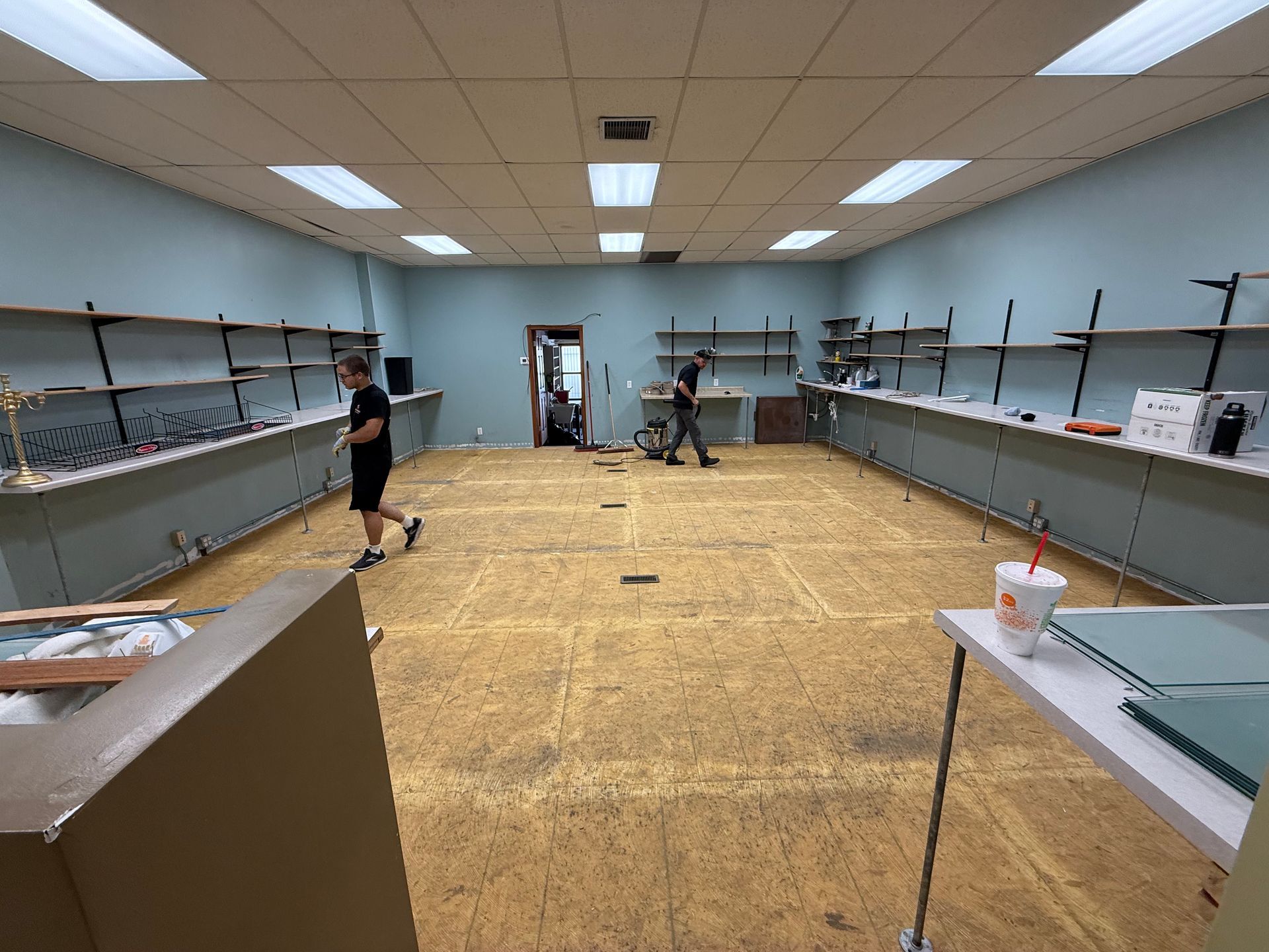 Empty classroom with shelves along both walls and two people working near the far end of the room.