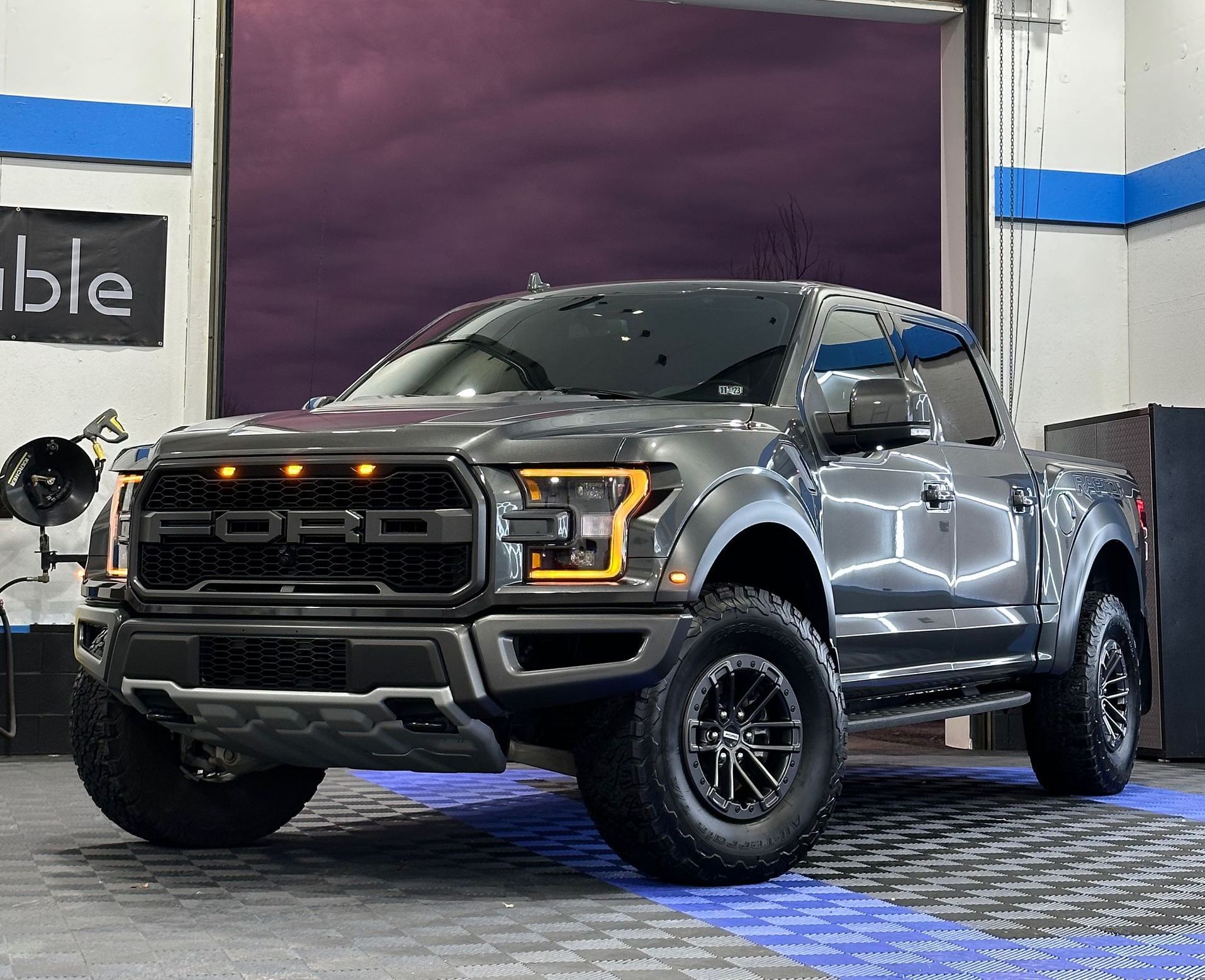 A gray Ford F-150 Raptor pickup truck parked inside a garage with bright lights on a checkered floor.