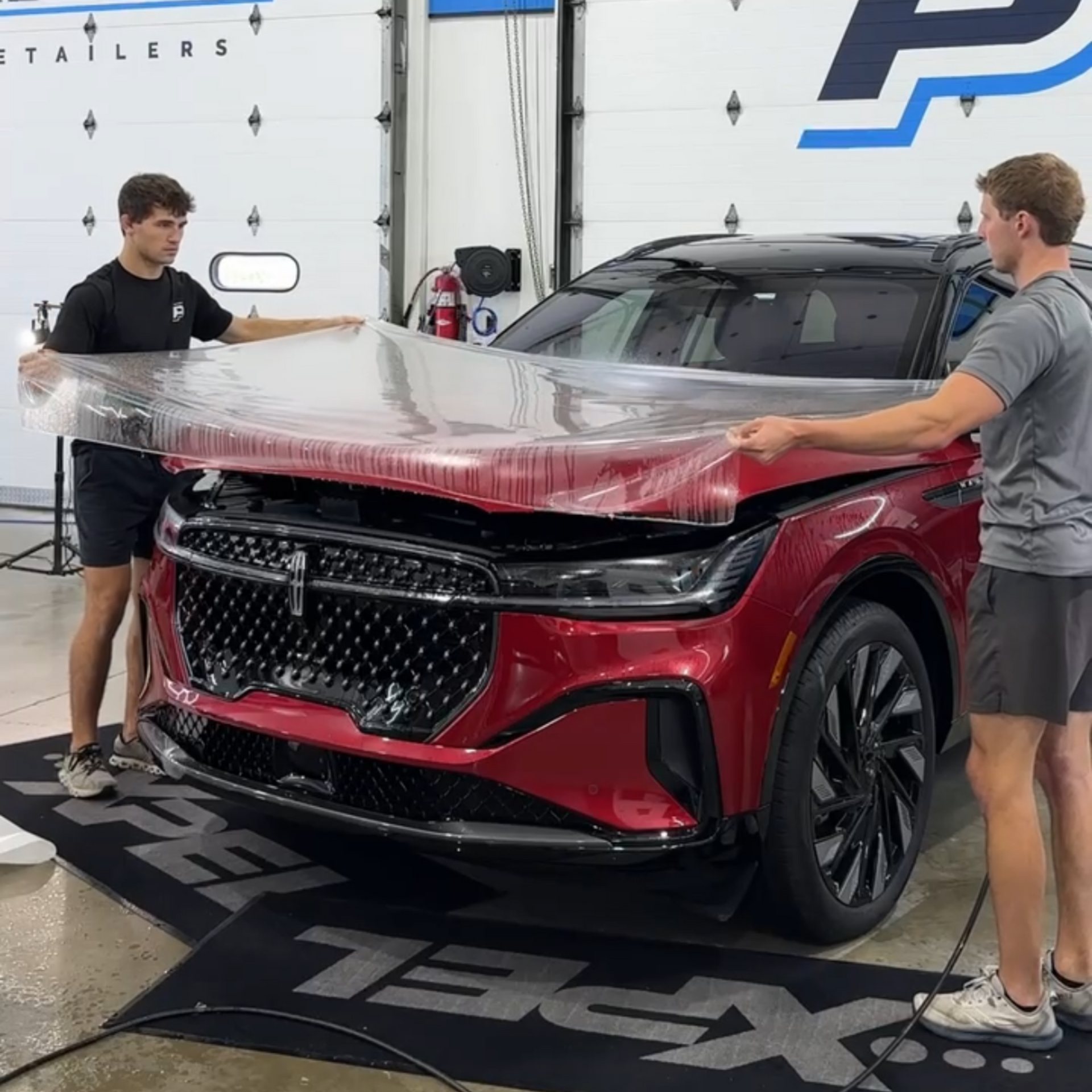 Two people working together to apply a clear paint protection film to the hood of a red SUV in a garage.