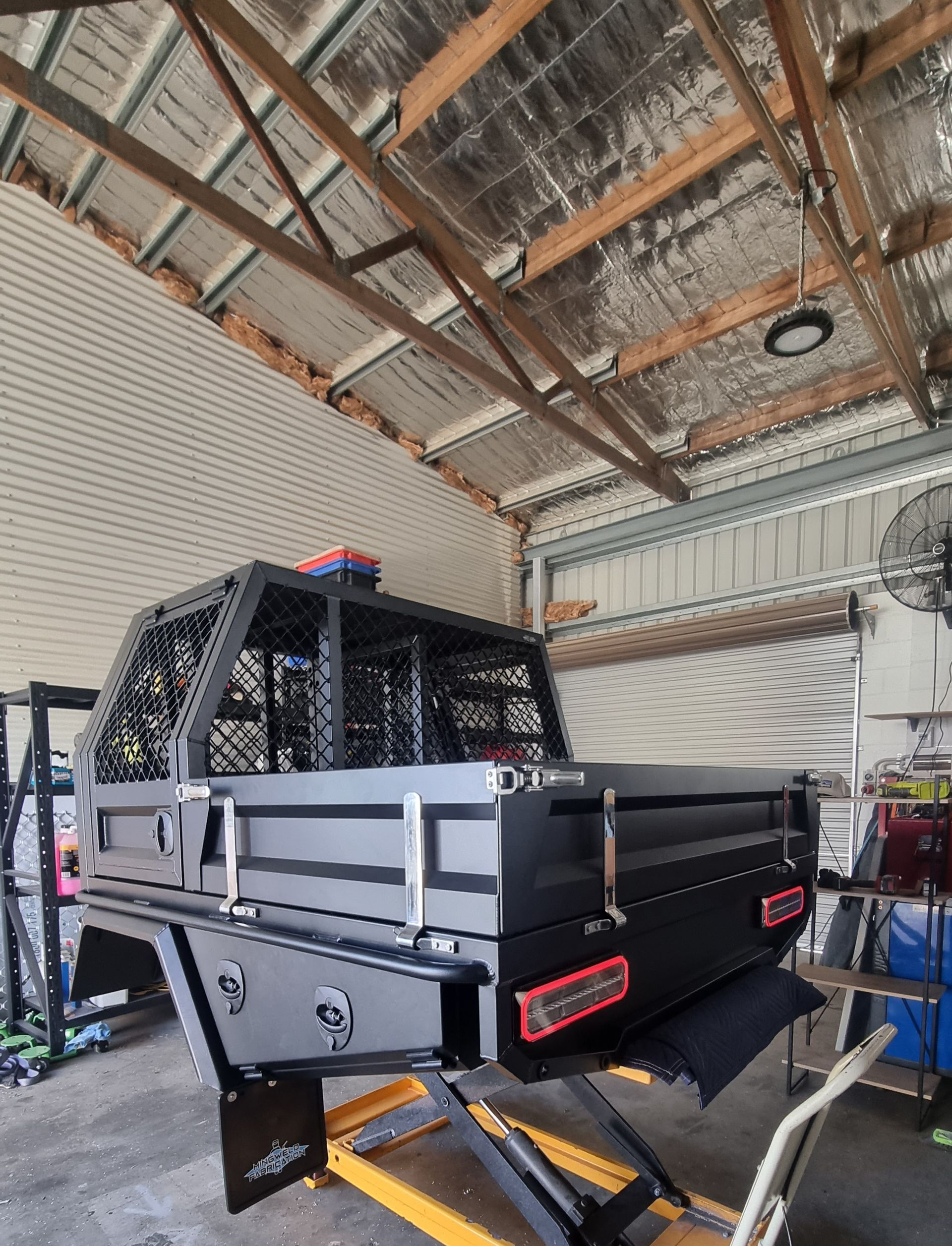 A black custom metal truck tray and canopy structure mounted on a yellow hydraulic lift in a workshop.