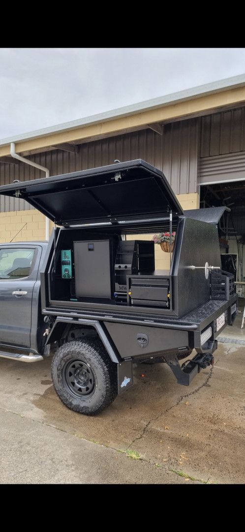 A matte black utility canopy mounted on a pickup truck, with its door open to reveal a built-in kitchen and fridge system.