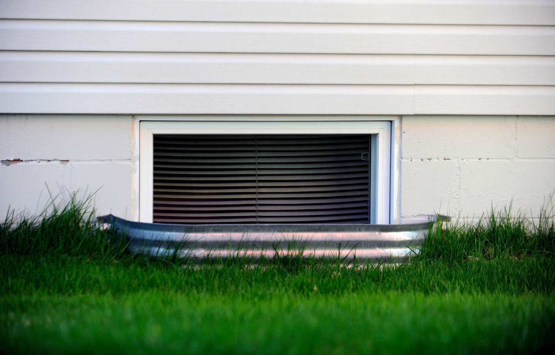 Basement window with black vent, centered in a white siding wall above green grass