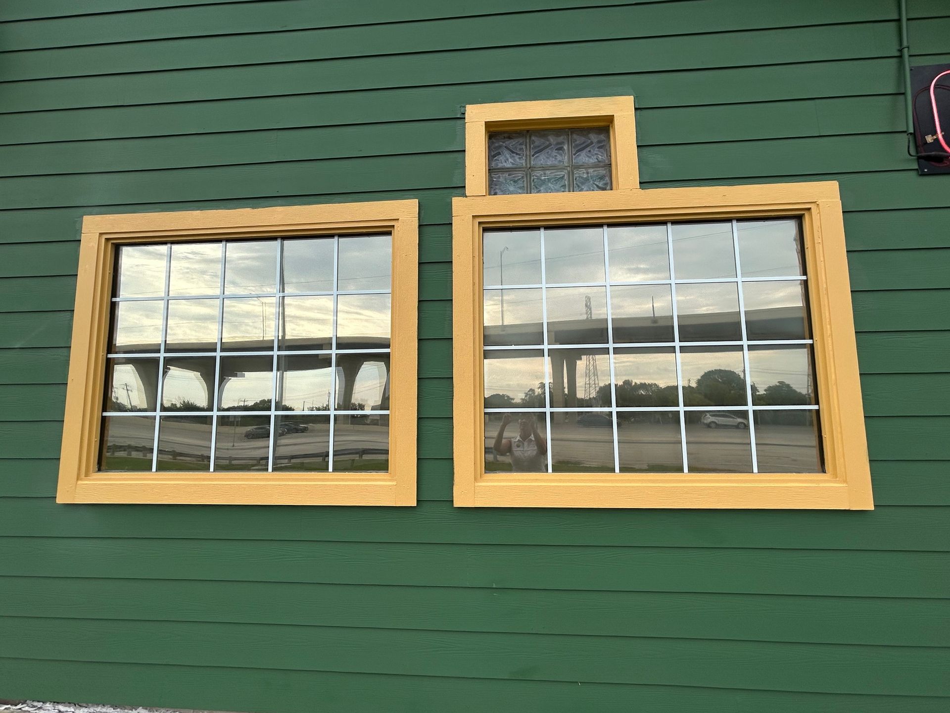 Three windows with yellow frames on a green building; a bridge is reflected in the glass.