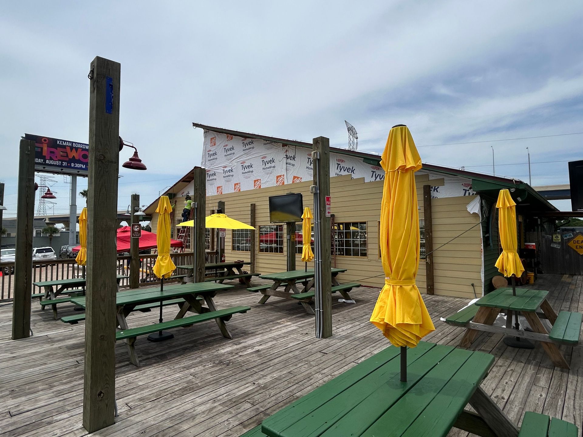Outdoor restaurant patio with yellow umbrellas, green picnic tables, and unfinished building siding.