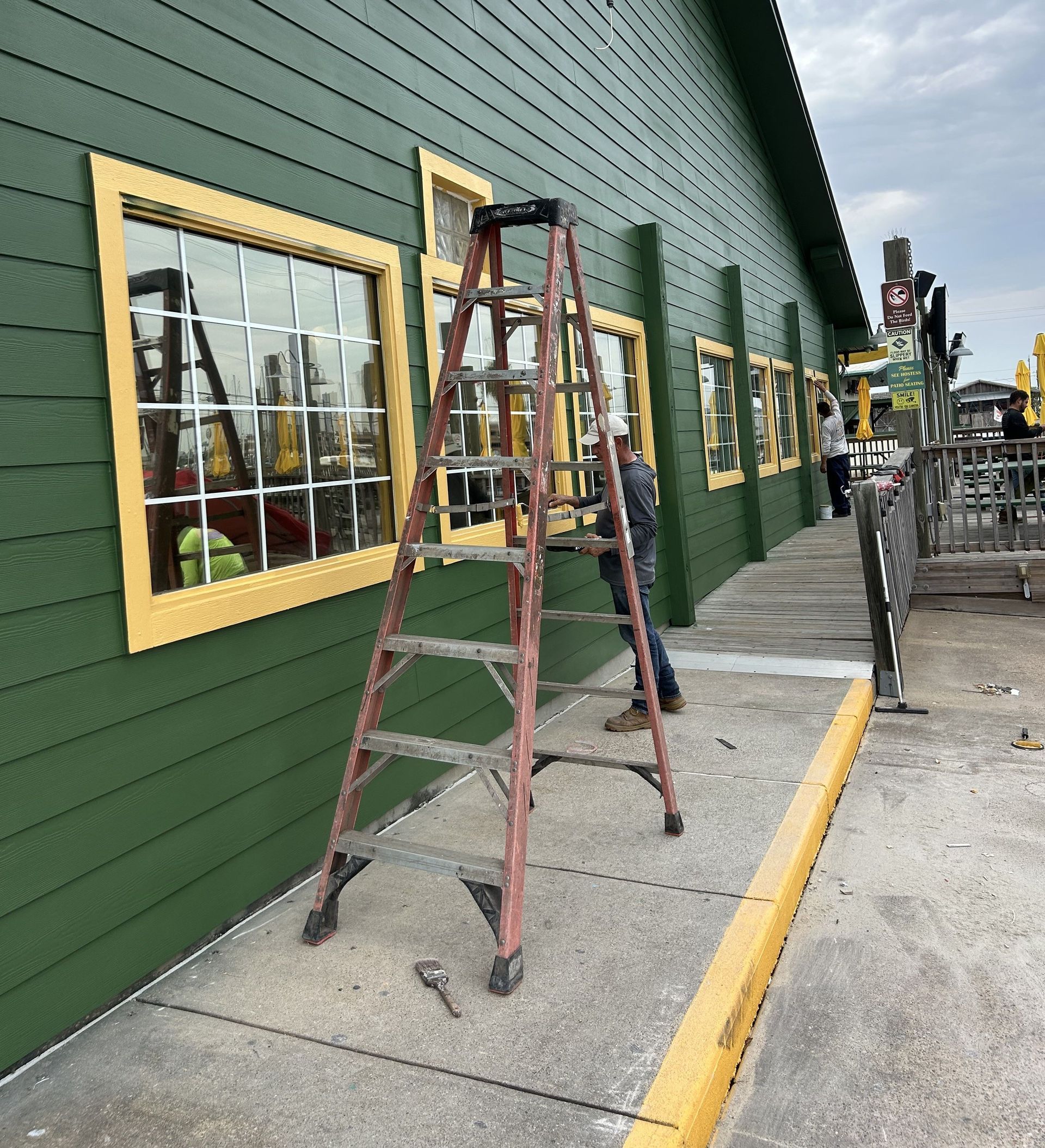 Person on ladder working on green building's windows with yellow trim. Concrete sidewalk with a yellow curb.