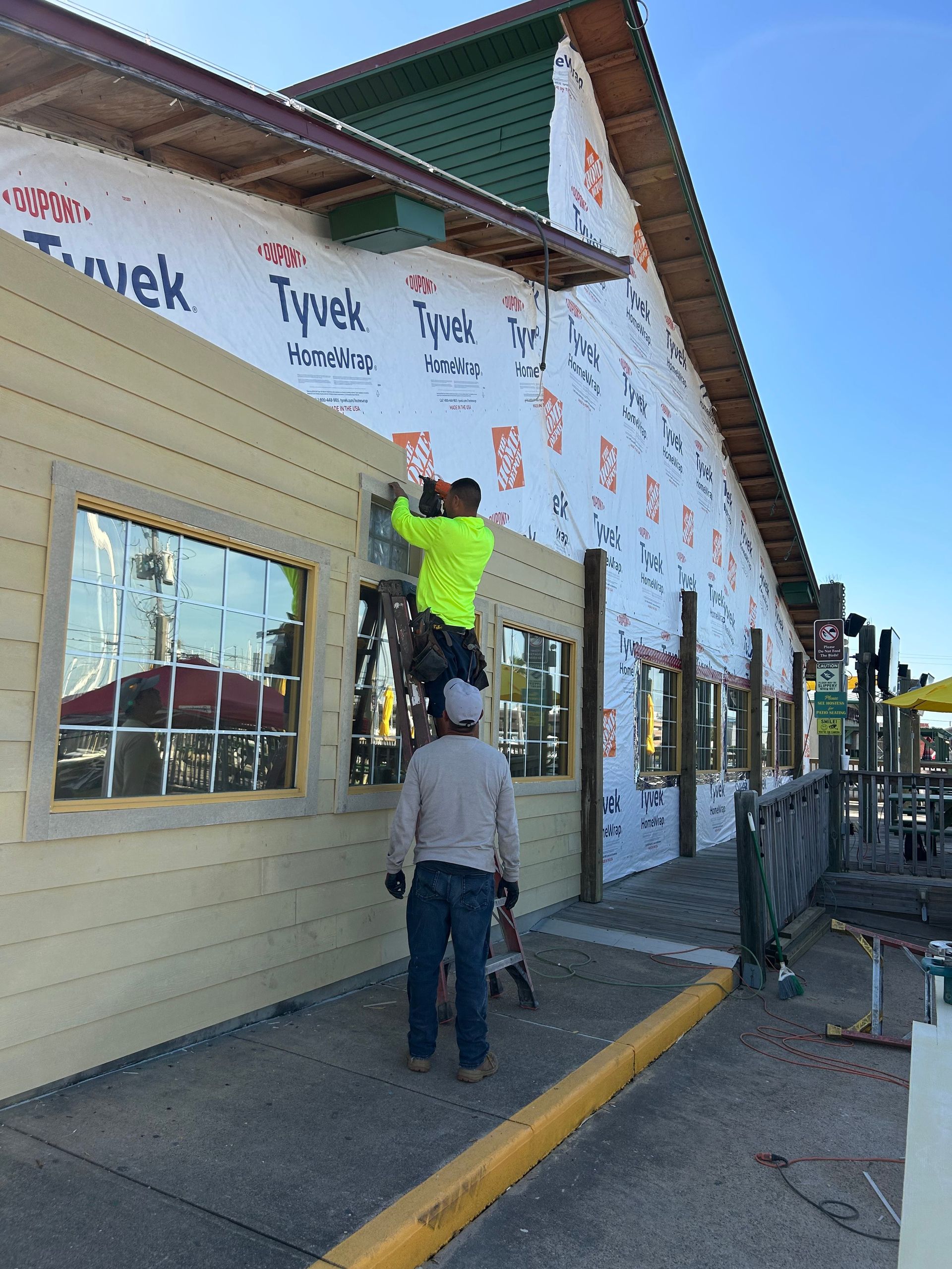 Construction workers installing siding on a building. One is on a ladder, wearing a neon vest. The building is wrapped in Tyvek.