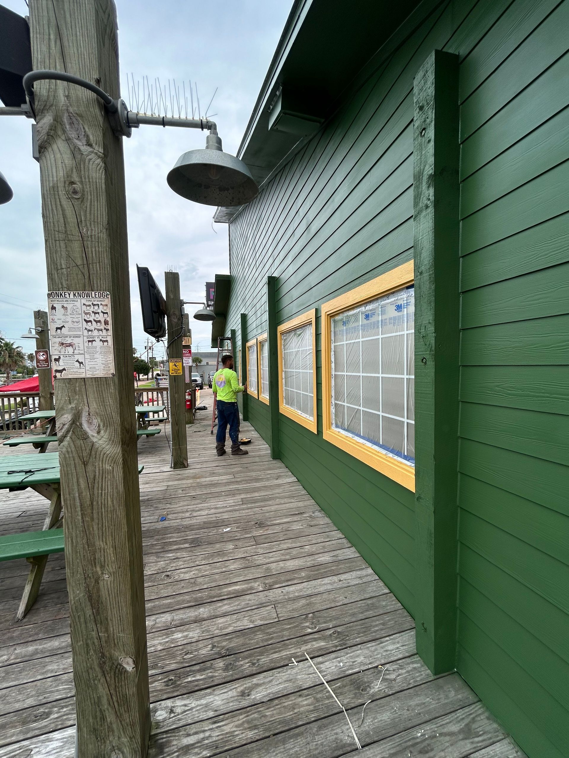 A person paints the exterior of a green building with yellow-trimmed windows on a boardwalk.