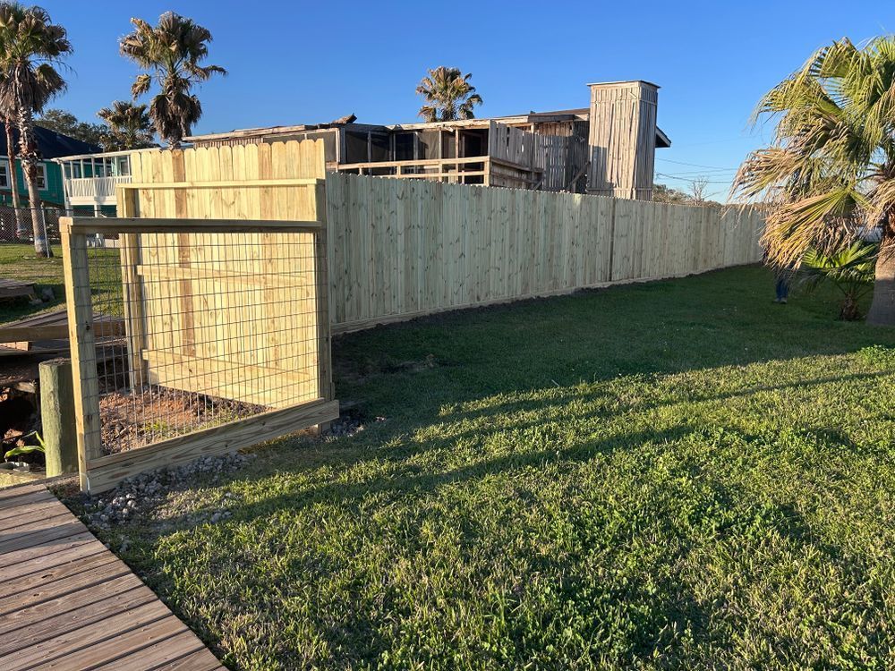 Wooden fence with gate surrounding a grassy yard, with a house and palm trees in the background.