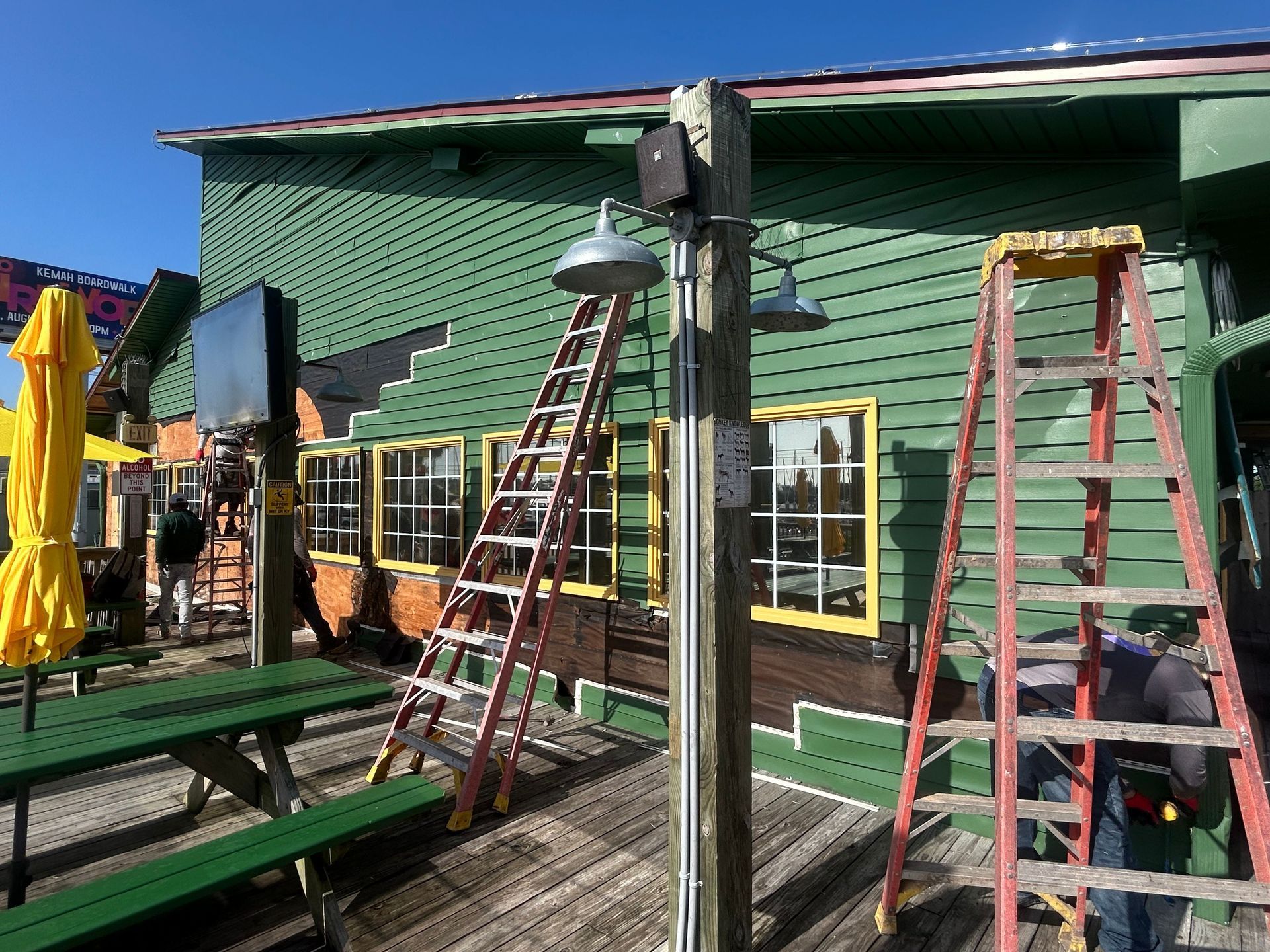 Two ladders leaned against a green building with yellow-framed windows. A person works on the building exterior.