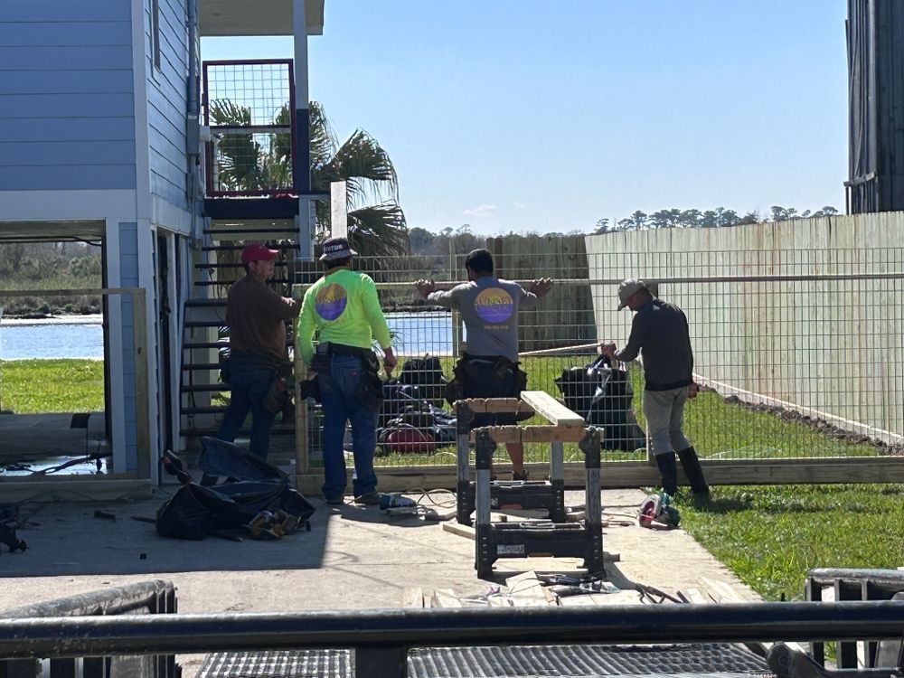 Construction workers building a fence near a light blue house and water on a sunny day.