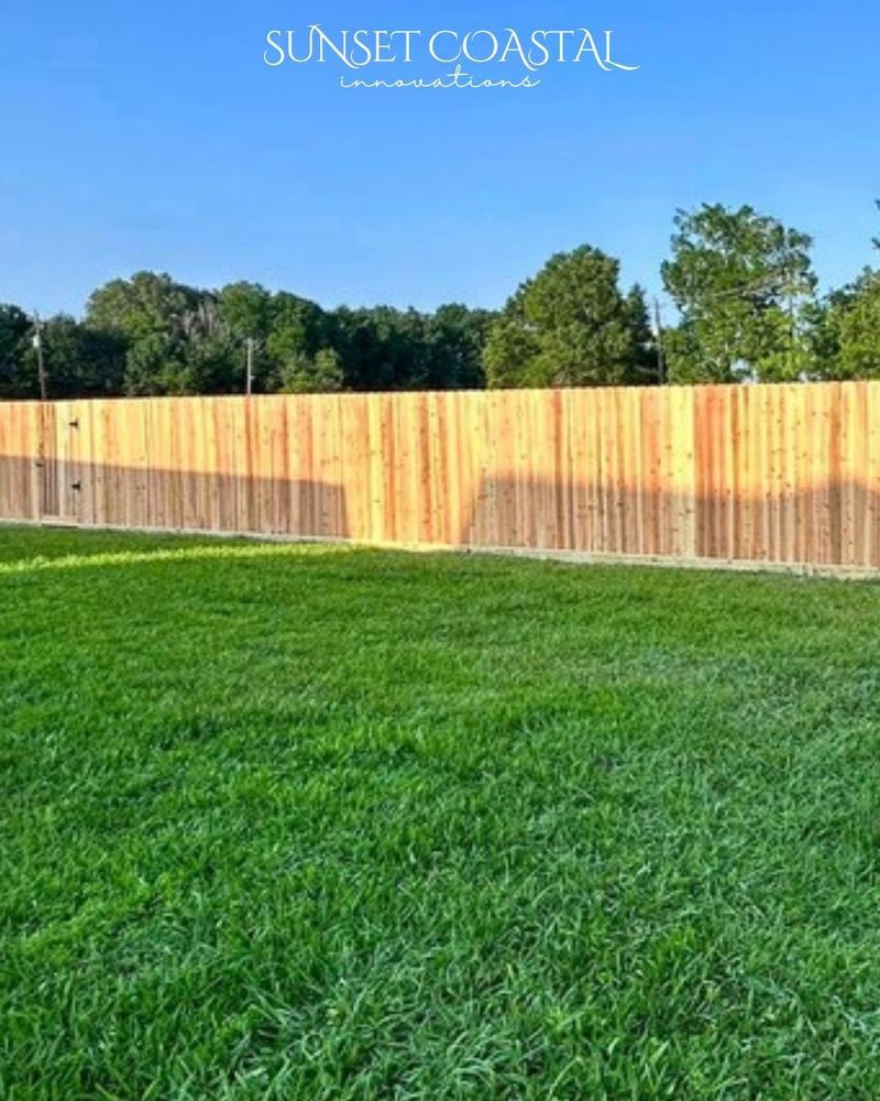 A wooden fence in front of green grass and trees under a clear blue sky.