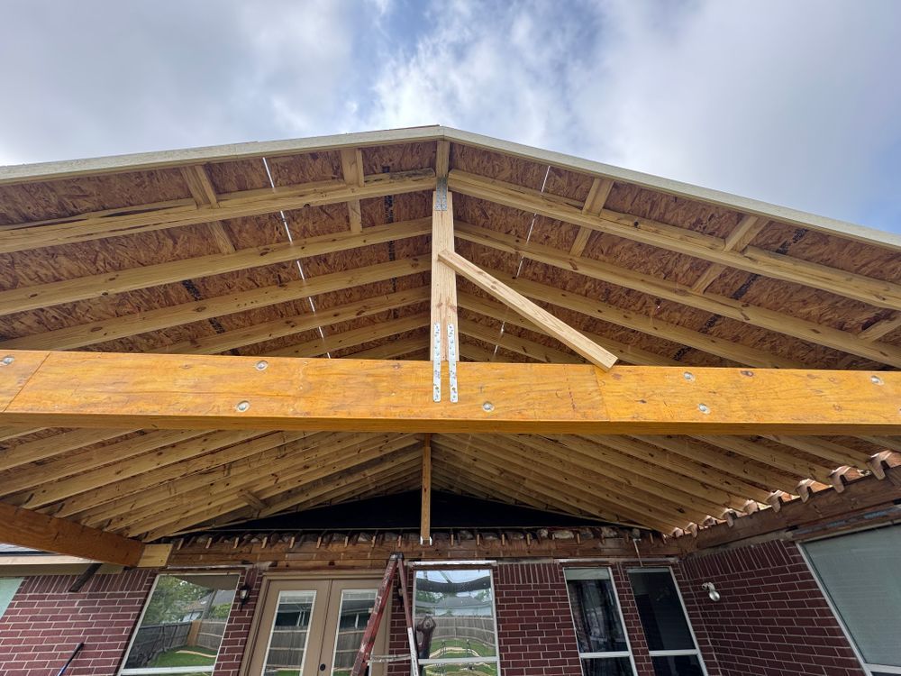 Wooden patio roof under construction, attached to a brick house with windows. Cloudy sky visible.