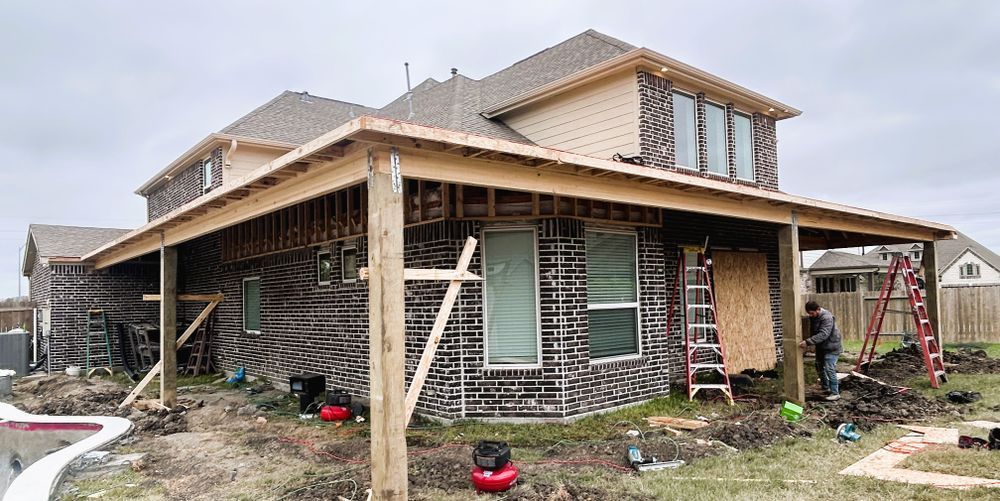 Construction of a wooden porch attached to a two-story house. A worker stands near a ladder. Overcast day.