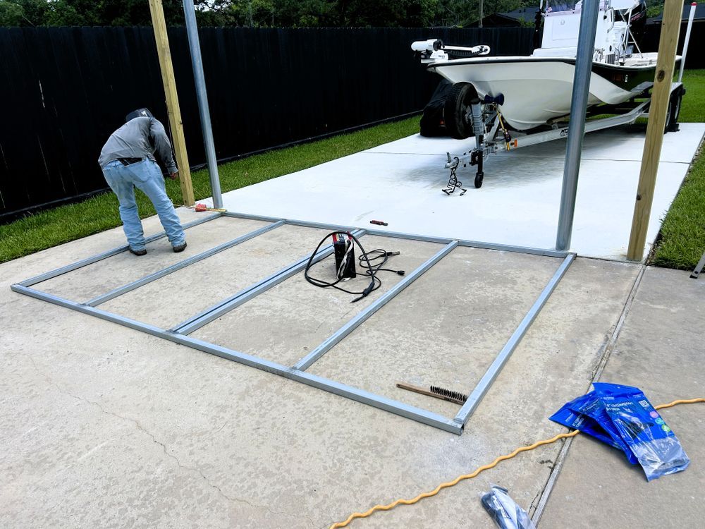 Person assembles metal frame carport base on a concrete driveway, a boat on trailer in the background.