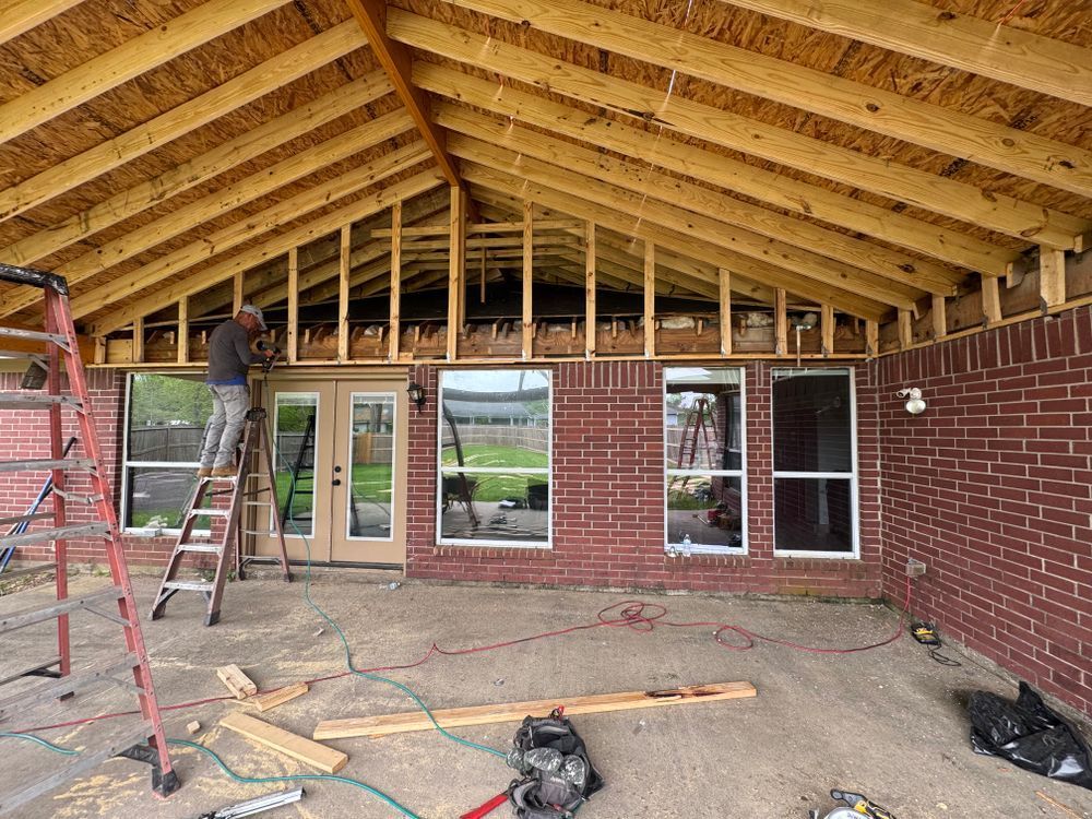 Construction of an outdoor patio cover. A worker on a ladder adds framing to a brick building's exterior.