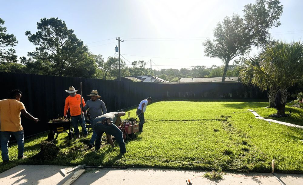 A crew of landscapers working in a sunny backyard. Some are using a tiller, and others are moving debris.