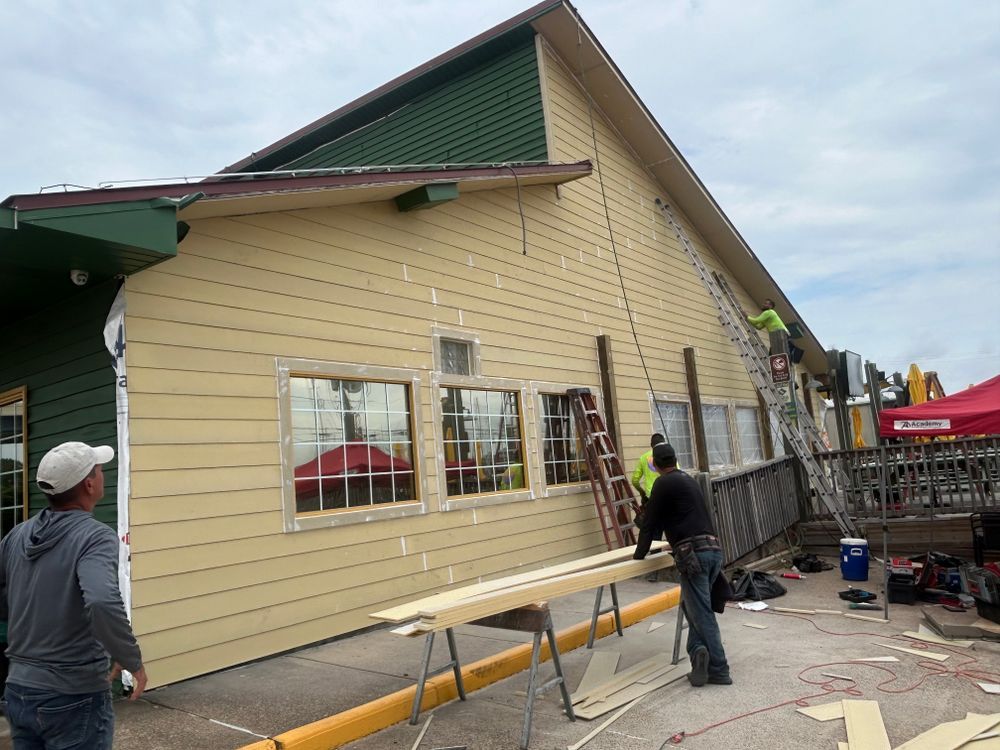 Workers installing siding on a tan building with green trim. Construction tools are present.