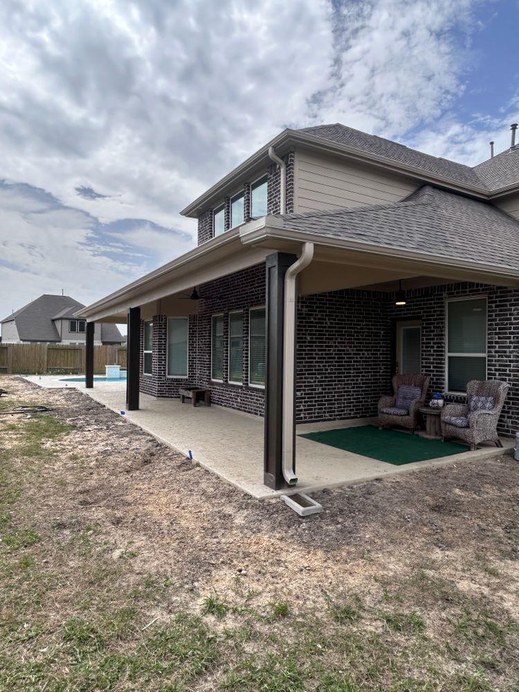 A covered patio attached to a brick house with outdoor seating, overlooking a pool and yard on a cloudy day.