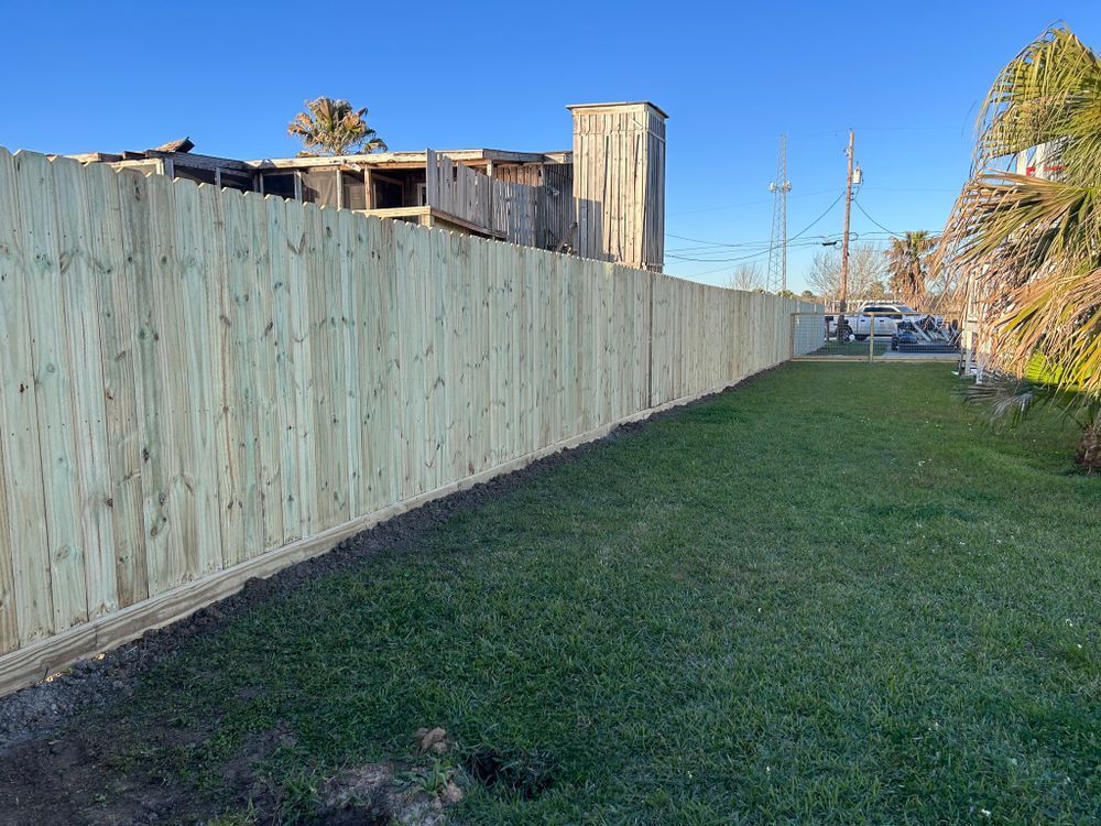 Wooden fence along a grassy area, with a partially visible building in the background under a blue sky.