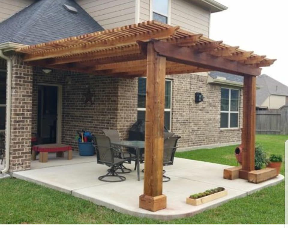 Wooden pergola over a concrete patio with a dining table, on a lawn next to a brick house.