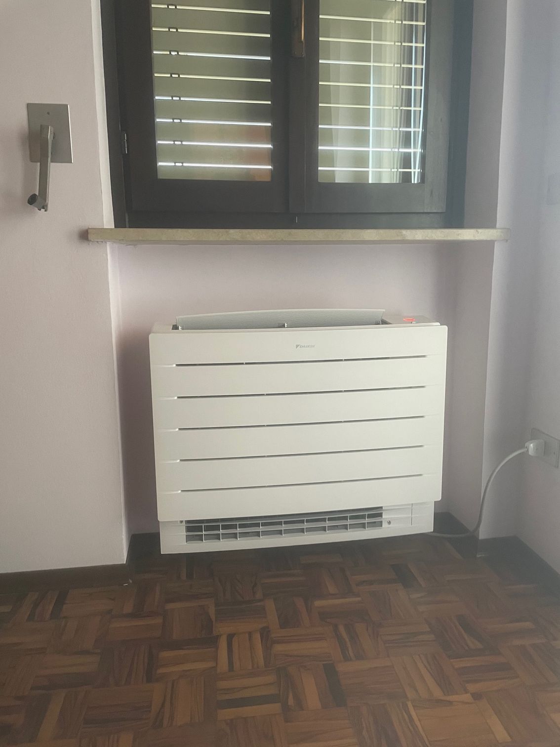 Un termosifone bianco sotto una finestra con le persiane chiuse, contro una parete rosa e un pavimento in legno.