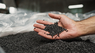 close-up of a hand releasing black recycled plastic granules into a pile, showing raw materials used in Brava composite roofing products.