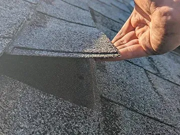 Hand lifting an asphalt roof shingle, revealing a gap, outdoors. Gray shingles and hand.