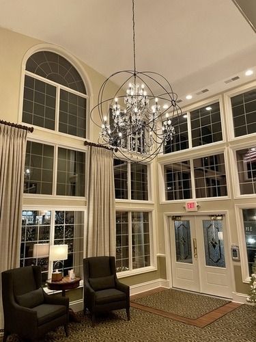 Elegant lobby with tall windows, chandelier, and two dark chairs by a table near glass doors.