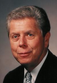 Portrait of a man in a dark suit and patterned tie, smiling against a gray studio background
