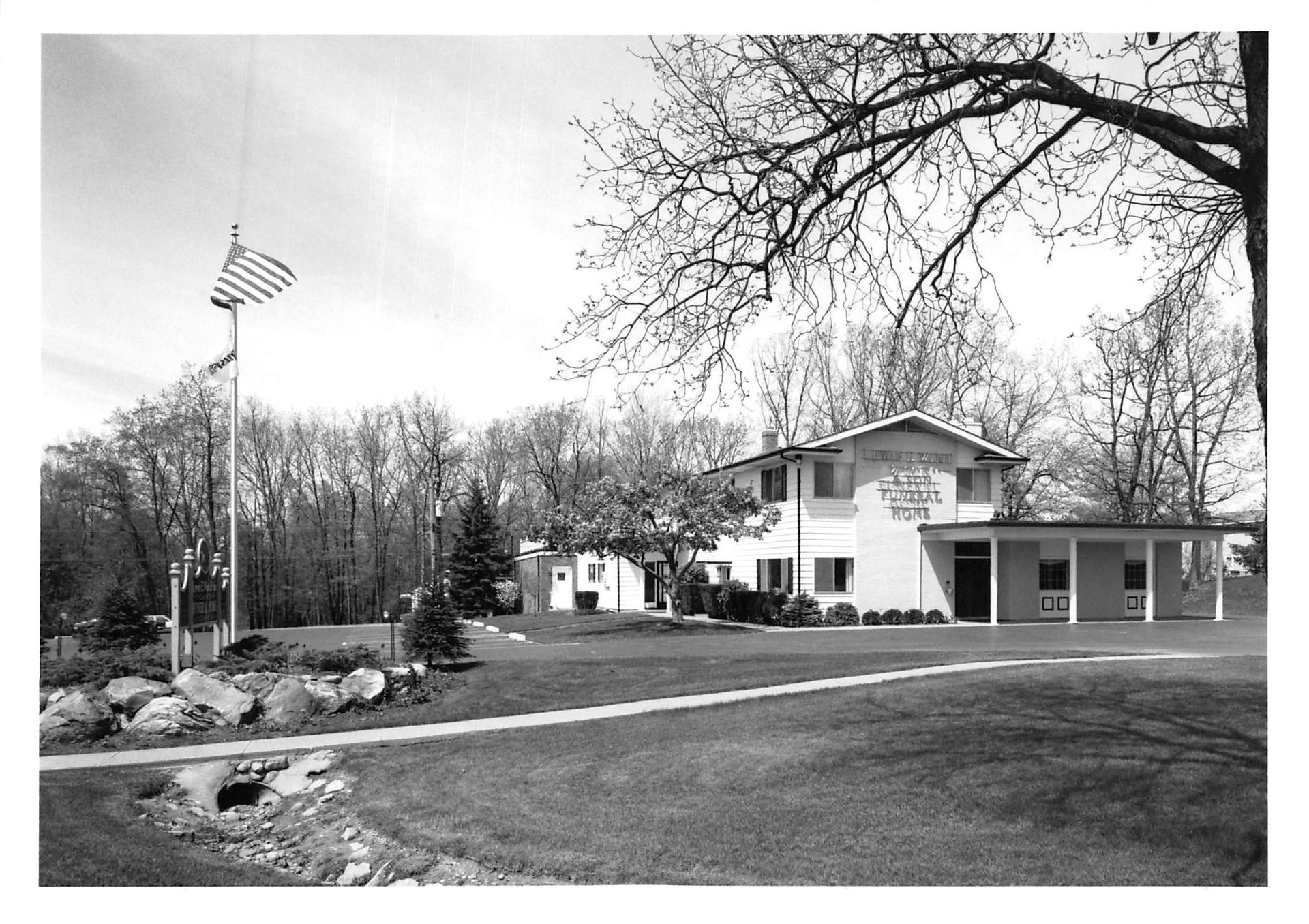 Black-and-white view of a house with an American flag, lawn, trees, and a garage under bare branches
