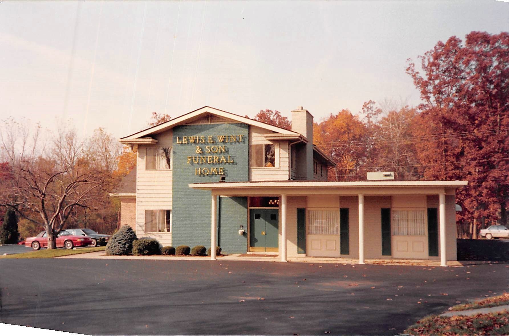 Small beige office building with covered entrance, green sign, and autumn trees beside a parking lot