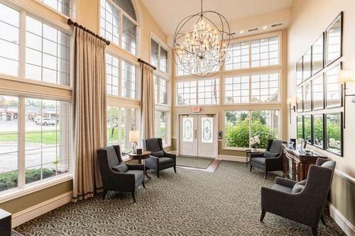 Bright sunroom lounge with tall windows, chandeliers, and dark chairs around a carpeted seating area