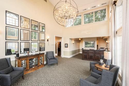 Bright lobby with framed wall art, gray chairs, and a dark wood reception desk under high windows.