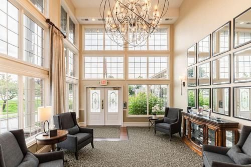 Bright hotel lobby with tall windows, chandeliers, armchairs, and a glass entrance door.
