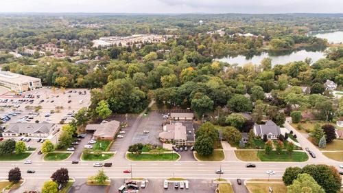 Aerial view of a suburban shopping center, parking lot, and surrounding trees with a pond in the distance