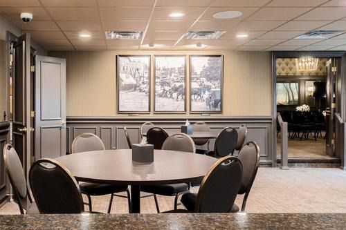 Empty lounge with round tables and chairs, framed artwork on beige walls, and an open doorway to a brighter room