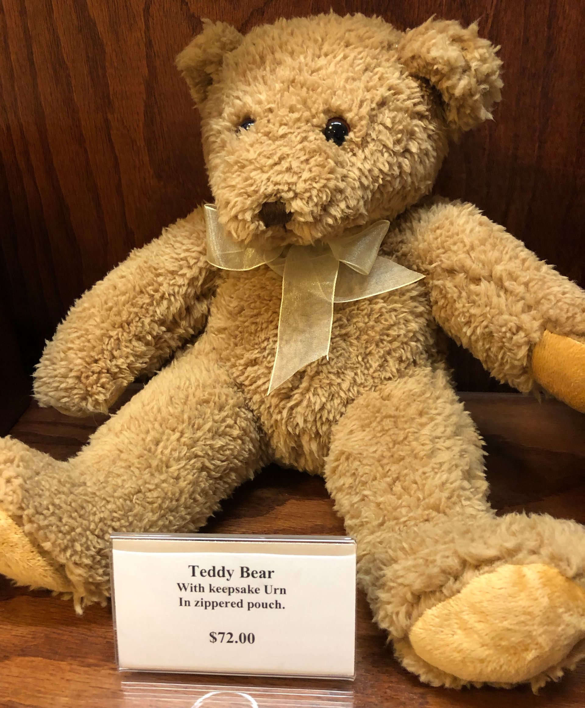 Tan teddy bear with ribbon bow, seated beside a price card on a wooden shelf.