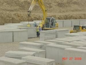Construction site with excavator lifting concrete blocks beside large stacked concrete slabs.