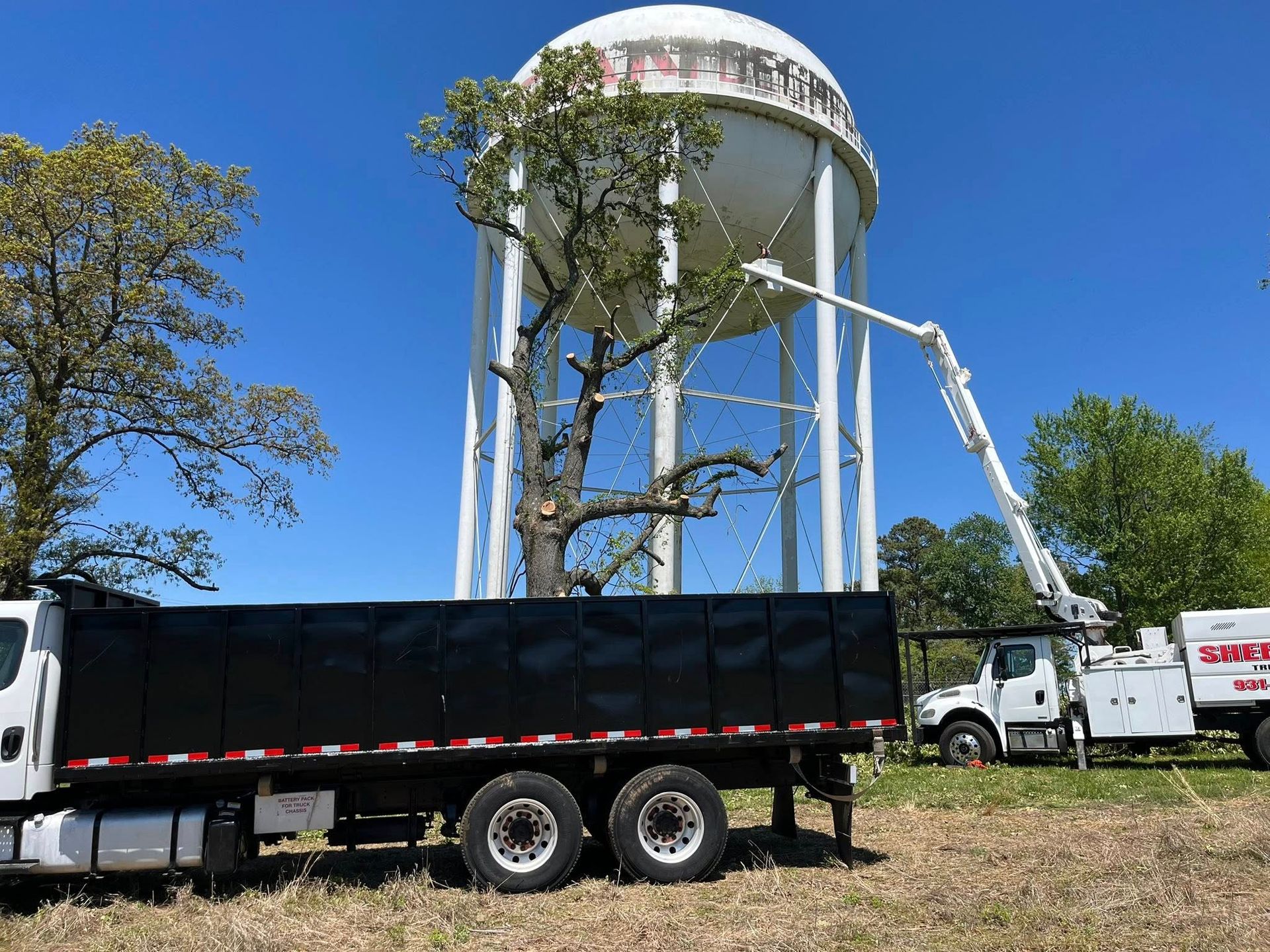 White water tower under repair with trucks and a crane in a grassy lot under a blue sky