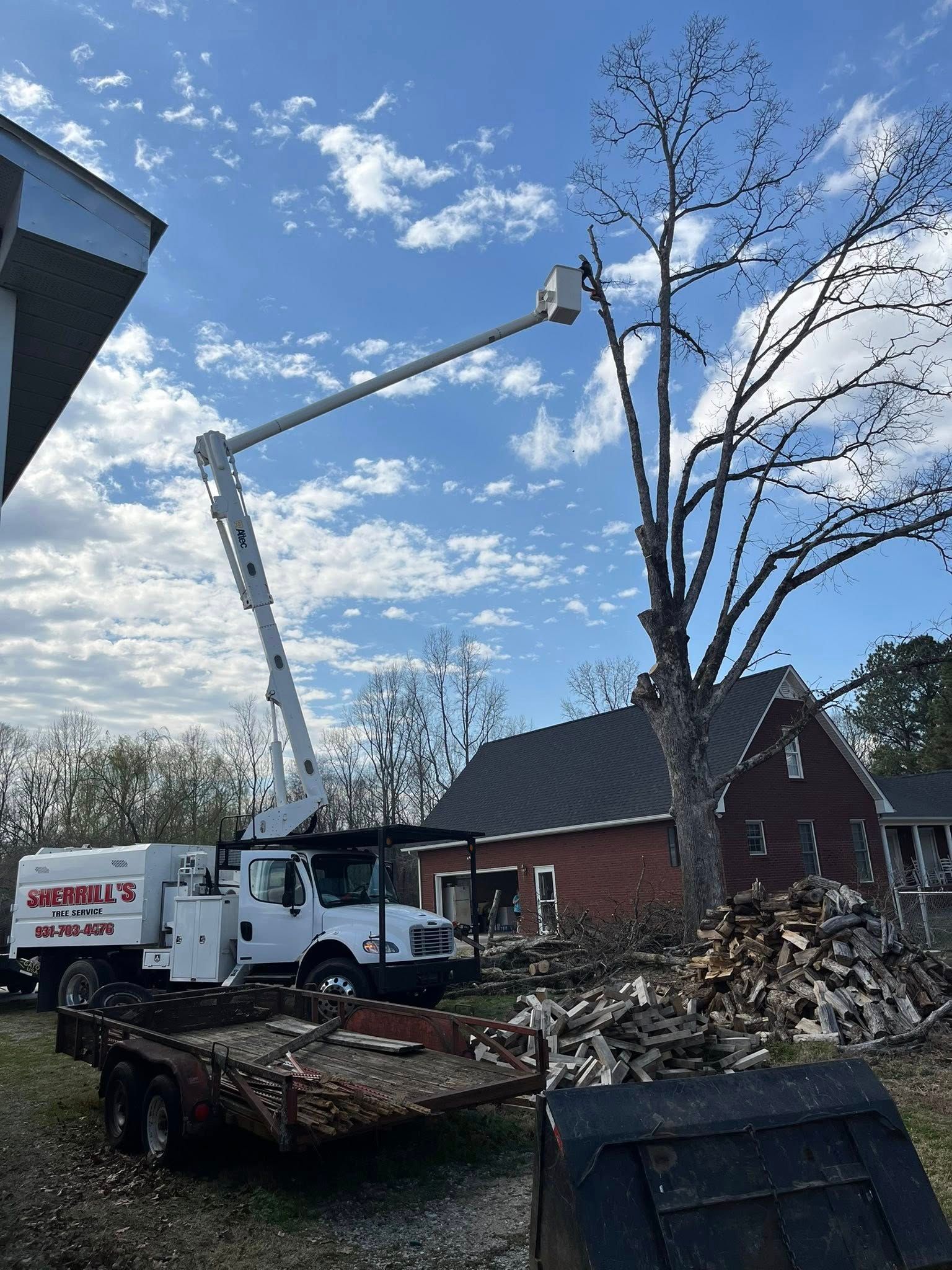 Construction truck lifting debris beside a house and tall tree under a blue sky