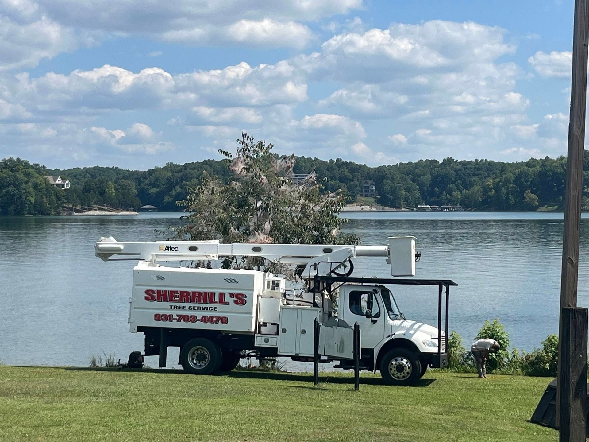 White utility truck parked by a lake with workers nearby and a boom arm, under a cloudy sky