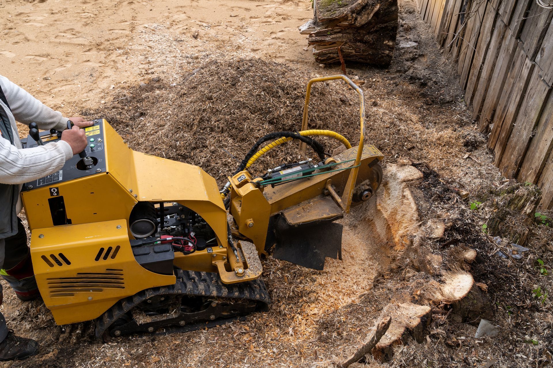 Yellow wood chipper grinding debris beside a fence, with a person operating it in a dirt yard.