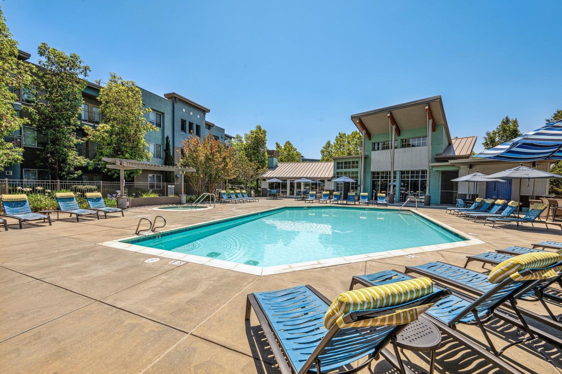 Swimming pool surrounded by lounge chairs, trees, and a building under a blue sky.