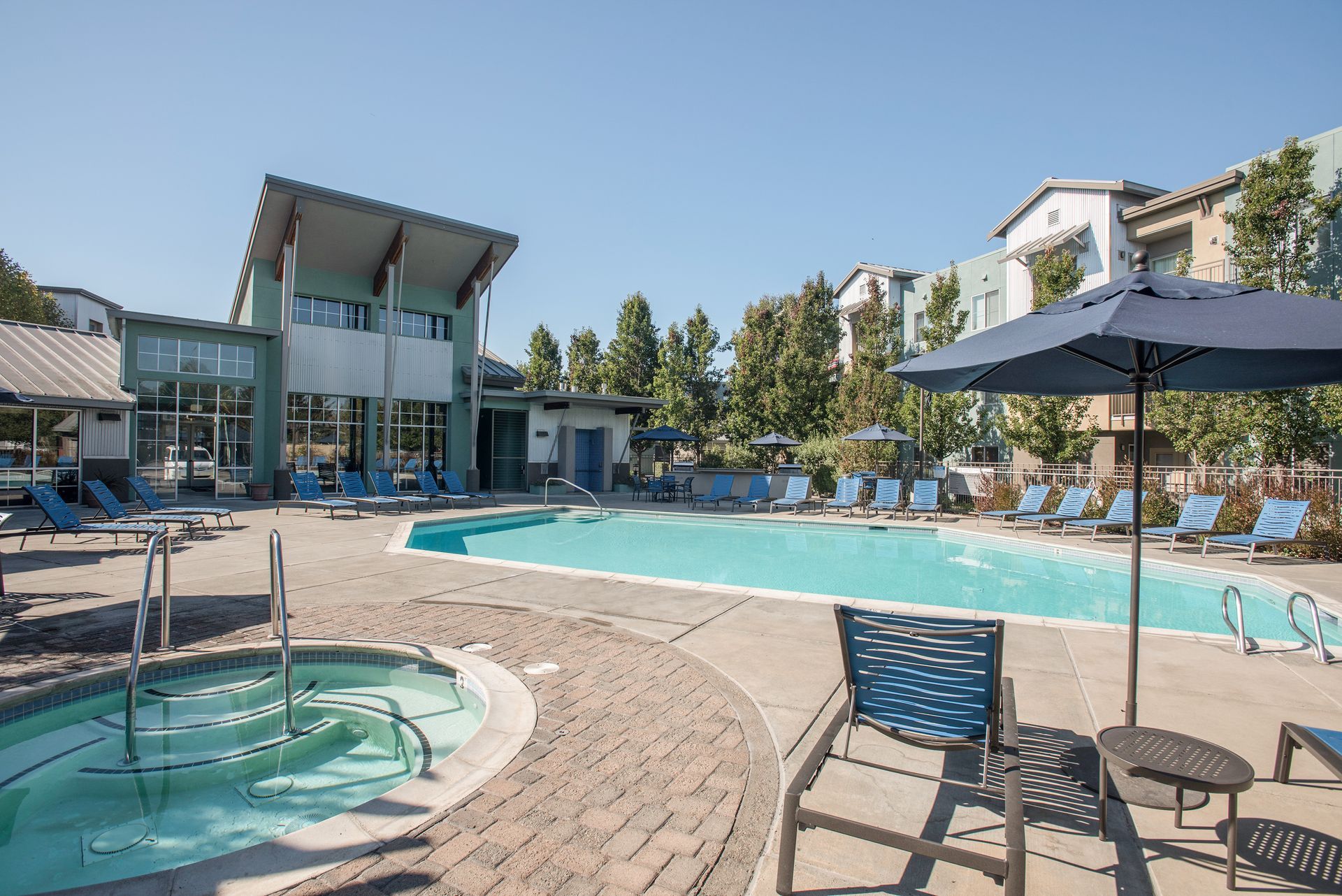 Pool and hot tub with lounge chairs and umbrella on a sunny day. Clubhouse in background.